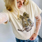 A graphic tee with a camo bow and boots design, predominantly in beige color, paired with blue denim shorts and white boots.