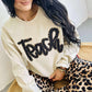Woman wearing a beige sweatshirt with 'Teach' in black sequins and leopard print pants sitting on a woven chair.