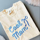 A yellow graphic tee shirt with blue embroidered lettering that reads 'Cool Mom' with a bow detail above the text.