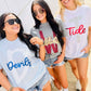 Three women wearing t-shirts with 'Devils', 'W', and 'Tide' glitter puff text, standing outdoors.