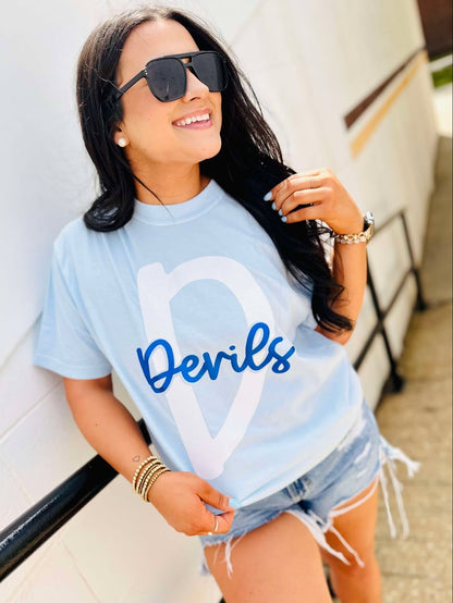 Woman wearing a light blue t-shirt with 'Devils' printed on it, standing against a white wall.