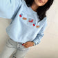 Person wearing a light blue sweatshirt with embroidered designs against a plain background