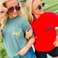Two women standing on a football field wearing matching graphic t-shirts. The woman on the left wears a muted sage green t-shirt with 'thornte' in gold script on the chest pocket, paired with denim shorts.