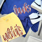 A flat-lay of two graphic t-shirts and a pair of white sneakers on a light gray tiled floor. The top blue t-shirt features 'hawks' in orange and white patterned script, and the bottom yellow t-shirt features 'wildcats' in a matching orange and white patterned script.