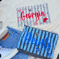 A flat-lay of a white graphic T-shirt with a Georgia Bulldogs sports design, styled with denim shorts, a matching blue striped sweatshirt, and white sneakers on a gray wood plank surface.