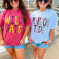 Two women standing outdoors in front of stadium seating, wearing oversized graphic t-shirts. The woman on the left wears a pink t-shirt with 'WILD CATS' in yellow and purple.