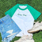 A styled flat-lay of a green and white raglan t-shirt with 'Game Day' printed on the chest, paired with distressed denim shorts, beige sneakers, and arranged on a green turf background.