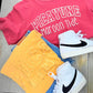 Pink t-shirt with 'PICAYUNE Maroon Tide', yellow towel, blue jeans, and white sneakers on a wooden floor.
