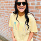 A person wearing a yellow graphic tee with the text 'MAMA' embroidered on the front, standing with one hand against a brick wall.