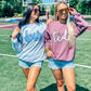 Two women standing on a field wearing sunglasses and custom mascot sweatshirts in blue and gray, with one woman's shirt displaying the word 'DEVILS' and the other's shirt displaying the word 'TIDE'.