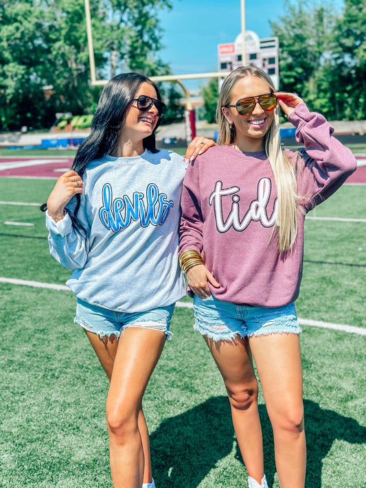 Two women standing on a field wearing sunglasses and custom mascot sweatshirts in blue and gray, with one woman's shirt displaying the word 'DEVILS' and the other's shirt displaying the word 'TIDE'.