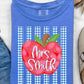 A folded blue T-shirt with a red apple graphic and the text 'Mrs. Smith' over blue-and-white gingham stripes, styled in a flat lay with jeans, sunglasses, a necklace, a slip-on shoe, and dried palm leaves on a white textured background.