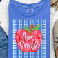 A folded blue T-shirt with a red apple graphic and the text 'Mrs. Smith' over blue-and-white gingham stripes, styled in a flat lay with jeans, sunglasses, a necklace, a slip-on shoe, and dried palm leaves on a white textured background.