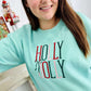 Woman wearing a light blue sweatshirt with 'Holly Jolly' thread, standing indoors.