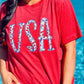 A person wearing a red t-shirt with the text 'USA' and an embroidered design resembling a flag on it, standing by a pool.