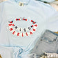 A light blue t-shirt with the text 'MISS AMERICA' and decorative friendship bracelet elements printed across the front, displayed flat with denim shorts and white sneakers.