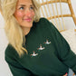 A woman sitting on a wooden chair wearing a green embroidered sweatshirt with a trio of mallard ducks on the front.