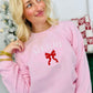 A woman wearing a light pink sweatshirt with 'Merry' embroidered on the front and a red bow detail.