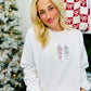 A woman sitting and wearing a white embroidered sweatshirt with the text 'Christmas Nutcracker Trio' on it, in front of a blurred Christmas tree and gifts in the background.