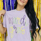 A person holding up a purple t-shirt with 'mardi gras' printed in yellow and neon green tinsel text.