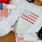 A sweatshirt with an embroidered American flag design in red and blue on a gray background, worn by a person seated in front of a small table decorated with American flags and related merchandise.