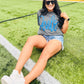 Woman sitting on a football field wearing a gray t-shirt with blue text and denim shorts.