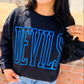 Person wearing a black sweatshirt with 'DEVILS' in blue letters against a brick wall.