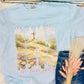 Light blue t-shirt with a printed design of a cross and flowers "He is not here. He has risen", held by a person wearing jeans.