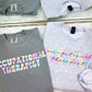 Gray t-shirts with pastel colorful 'occupational therapist' text on a mirror surface.