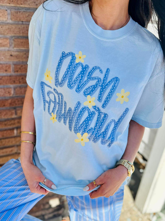 Person wearing a light blue t-shirt with 'Dash Forward' text and stars, standing against a brick wall.