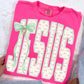 Pink t-shirt with 'Jesus' printed on it, featuring a green bow and cross charm, held by a hand.