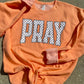 Orange sweatshirt with 'PRAY' printed on it in white letters with black polka dots, laid flat on a concrete surface.