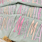 Stack of Bay Comfort Color tees with pink and white MAMA AUNTIE NONNI ABUELA NANNY LOLLI text on a light surface.
