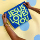 Blue pillow with 'Jesus Loves You' text held by a person against a polka dot background