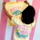 Person holding a yellow t-shirt with 'LATER' and an alligator graphic, wearing a white t-shirt with 'AFTER WHILE' and alligator graphic.