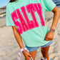 Person wearing a light green t-shirt with 'SALTY' in pink on a beach.