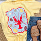 Yellow t-shirt with a red lobster design on a woven surface with jeans and sandals.