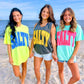 Three women on a beach wearing 'SALTY' t-shirts in different colors.