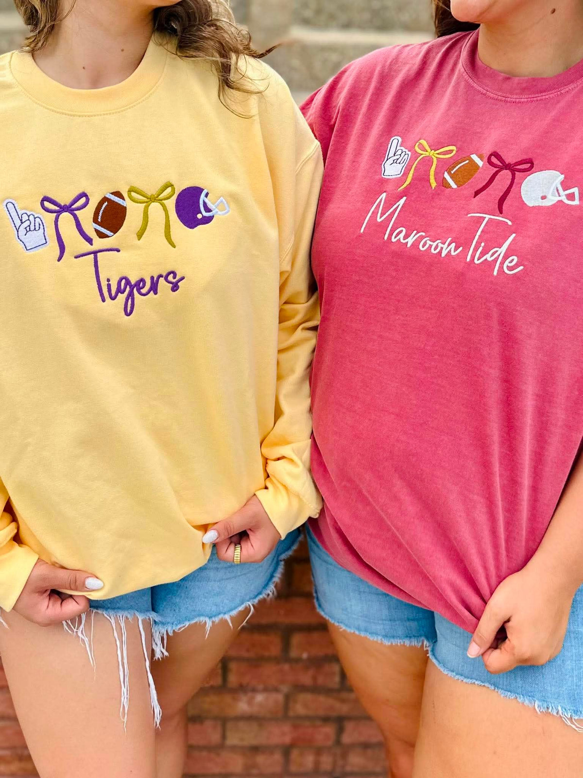 Two women wearing graphic crewneck sweatshirts, one in pale yellow with purple and gold football-themed graphics and the word 'Tigers', and the other in heather pink with white 'Maroon Tide' script and red, gold, and white graphics. They are wearing denim shorts against a brick wall background.