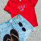 Red 'USA' t-shirt with denim shorts and sandals on a concrete surface