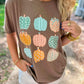 A woman wearing a brown short-sleeve t-shirt with a graphic print of colorful patterned pumpkins, photographed outdoors on a path.