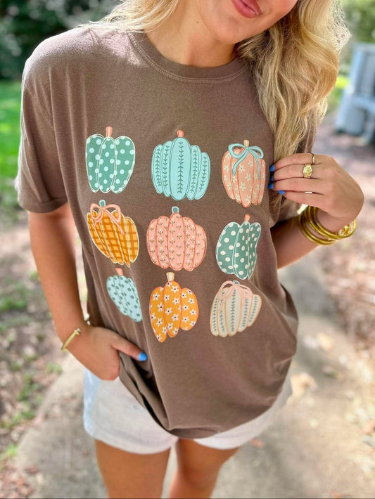 A woman wearing a brown short-sleeve t-shirt with a graphic print of colorful patterned pumpkins, photographed outdoors on a path.