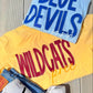 Yellow sweatshirt with 'WILDCATS' text, blue shirt with 'BLUE DEVILS' text, and white sneakers with black Nike logo on a wooden surface.
