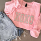 Pink sweatshirt with 'XOXO' Glitter Applique embroidery and denim shorts on a gray background