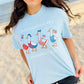 Light blue t-shirt with patriotic duck graphics worn by a person on a beach.