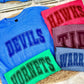 Collection of t-shirts with 'DEVILS', 'HAWKS', 'TIDE', and 'WARRIORS' text on a textured surface.