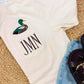 An off-white short-sleeve t-shirt with a front pocket featuring a duck graphic and the monogram 'JMN', styled in a flat lay with light-wash denim shorts and dark brown slide sandals on a woven rug.