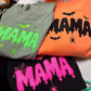 Three folded Halloween-themed t-shirts displayed together on a white surface. The top-left shirt is sage green with neon green dripping 'MAMA' text and small green spider/bat graphics.