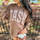 A woman wearing a brown sweatshirt with 'DASH' printed in large white and tan zebra-patterned letters, paired with patterned shorts and sunglasses, standing outdoors in front of a fence.