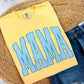 A folded yellow t-shirt with a blue striped 'MAMA' graphic, styled on a woven round mat with blue denim shorts.